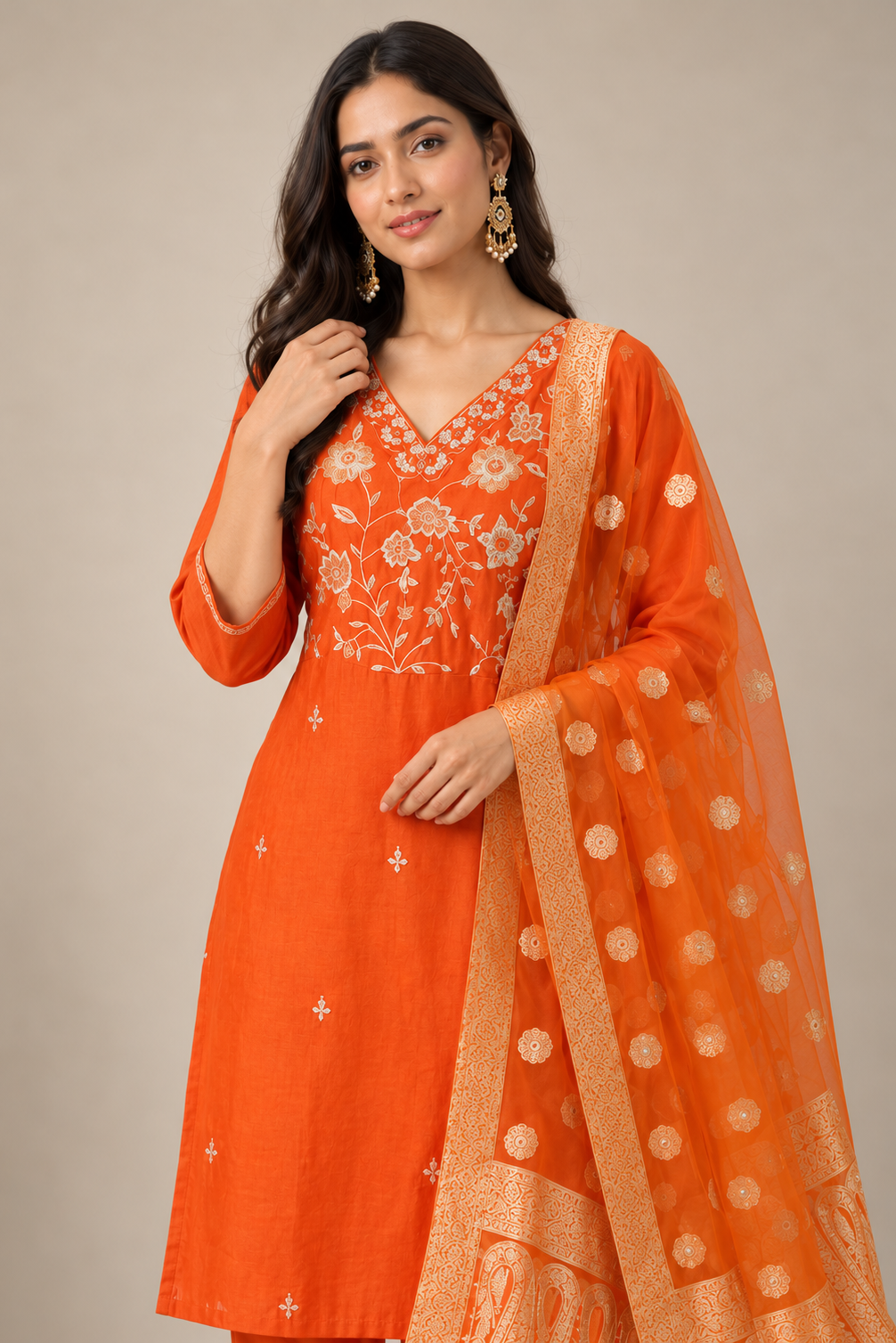 A woman modeling a bright orange traditional Indian outfit with white floral embroidery on the bodice, matching embroidered details on the kurta, and a sheer orange dupatta with gold zari motifs and border.