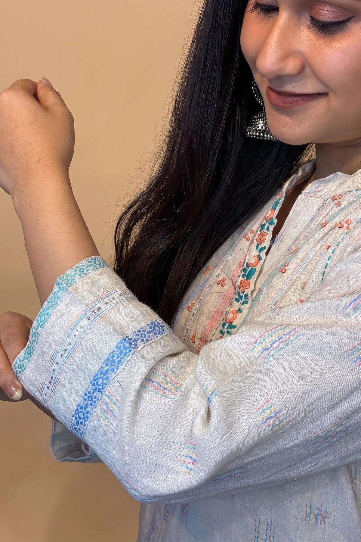A woman wearing a white embroidered kurta with blue patterned cuffs and a floral neckline, shown in a close-up shot against a plain beige background.