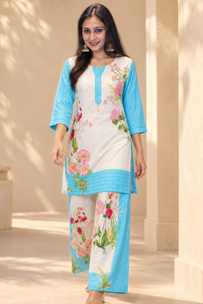 A woman modeling a traditional Indian three-piece salwar kameez set. The set features a cream-colored kurta with light blue sleeves and hem, decorated with pink and green floral prints, paired with matching light blue trousers.
