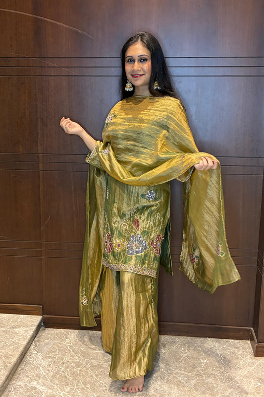 A woman modeling a metallic olive green traditional Indian outfit with floral embroidery, standing against a dark wood-paneled wall.