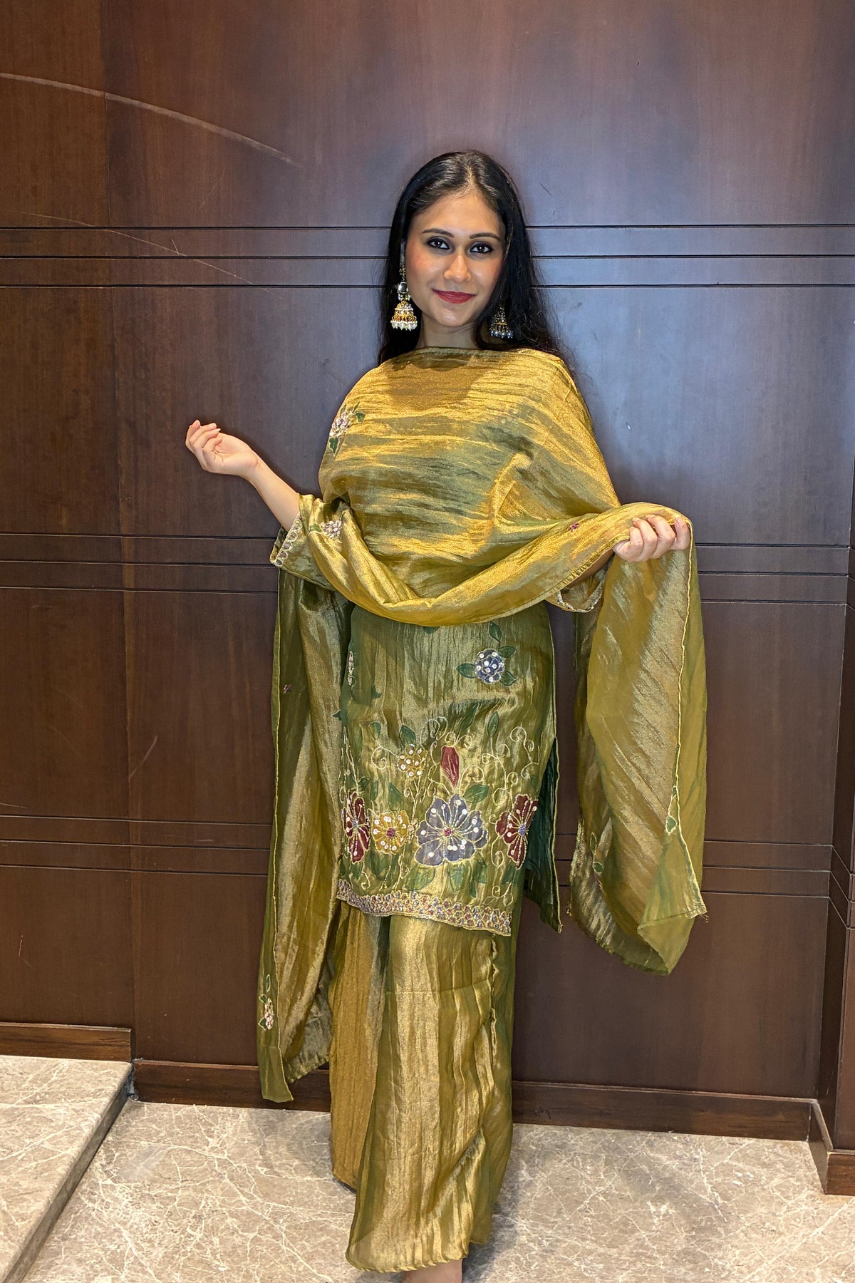 A woman modeling a metallic olive green traditional outfit with floral embroidery on the kurta and a matching dupatta, standing against a dark wood-paneled wall.