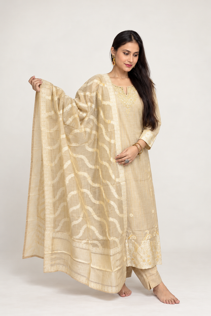 A woman modeling a traditional beige salwar kameez suit with a matching patterned dupatta, set against a plain light gray studio background.