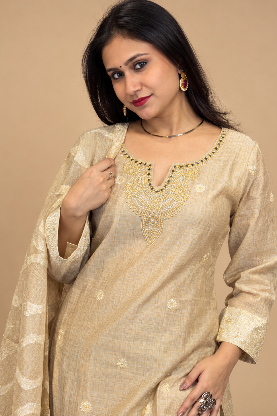 A woman modeling a beige traditional Indian salwar kameez suit with a matching dupatta. The kurta features gold and green embroidered detailing around the neckline, and she is posed against a solid beige background.