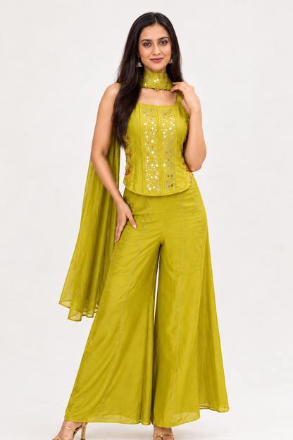 A woman modeling a bright yellow-green sleeveless traditional outfit with wide-leg pants, a matching choker, and a draped cape, set against a plain light gray studio background.