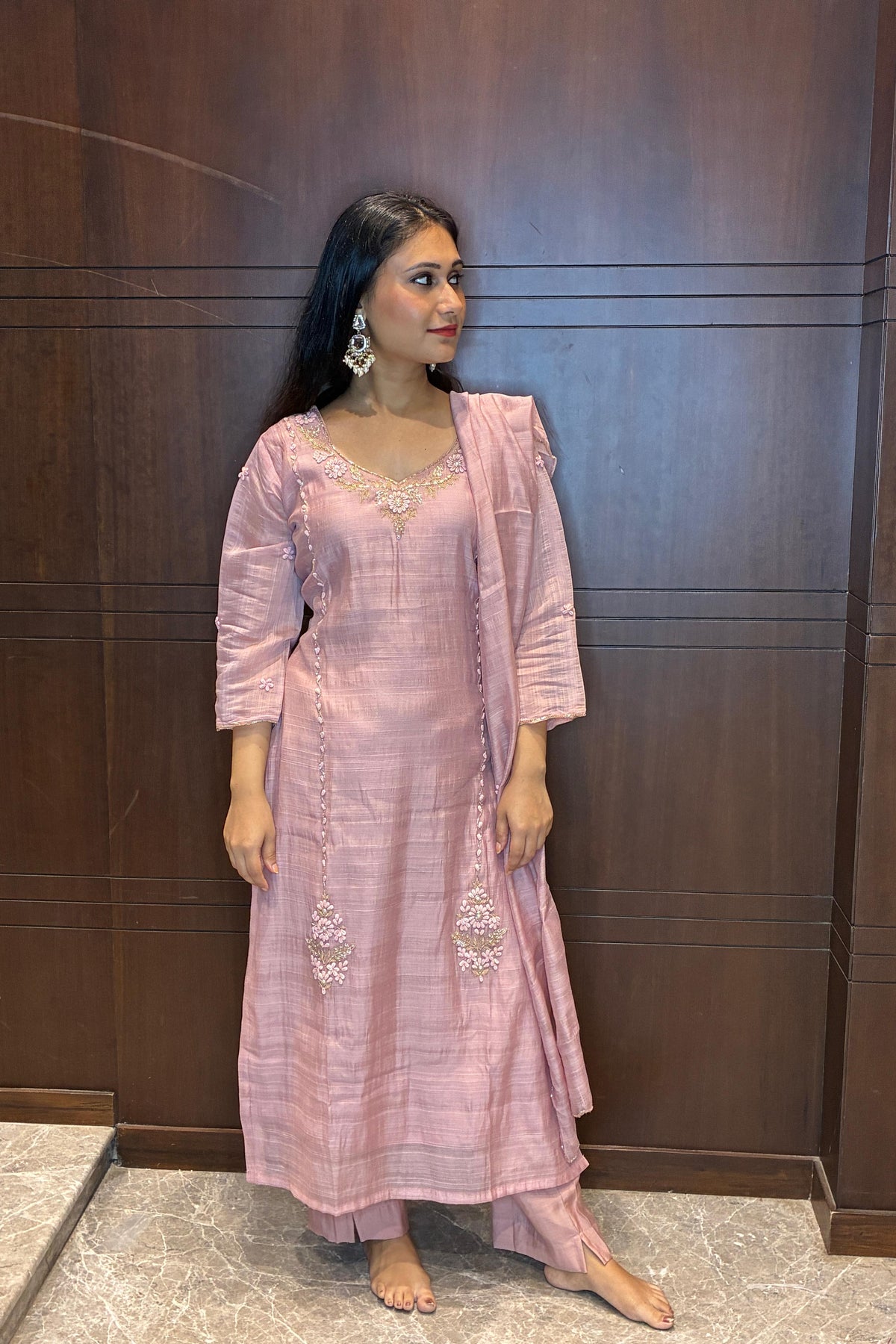 A woman modeling a light pink traditional Indian outfit with a matching dupatta, standing against a dark wood-paneled wall.