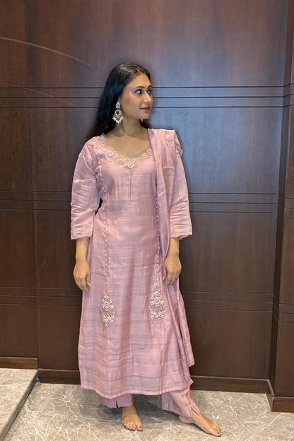 A woman modeling a light pink traditional Indian outfit with a matching dupatta, standing against a dark wood-paneled wall.