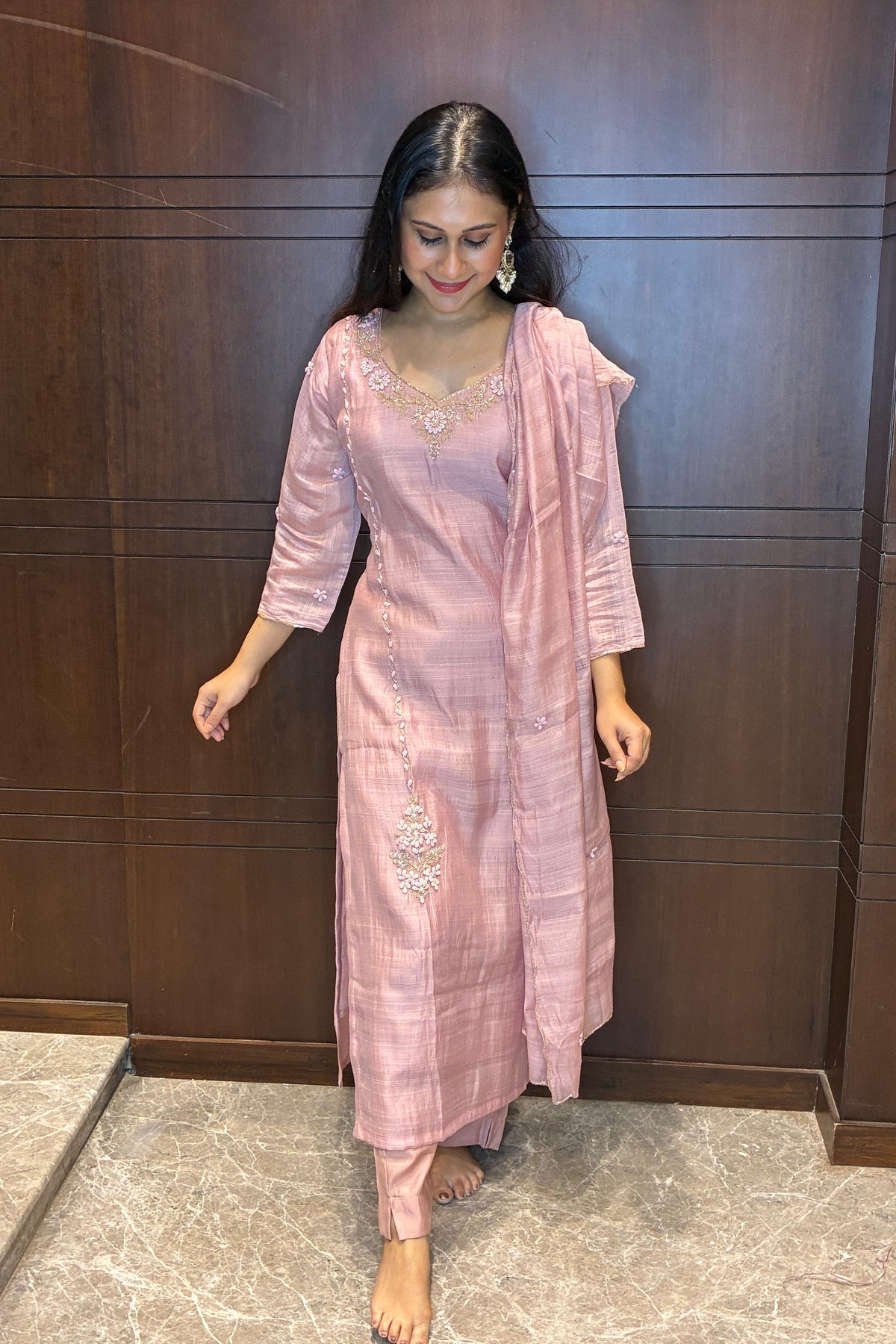 A woman wearing a light pink traditional Indian outfit with a matching dupatta, standing in front of a dark wood-paneled wall.
