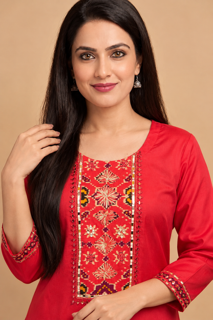 A woman with long dark hair wearing a bright red kurta with intricate multicolor embroidery on the chest and cuffs, posed against a solid beige background.