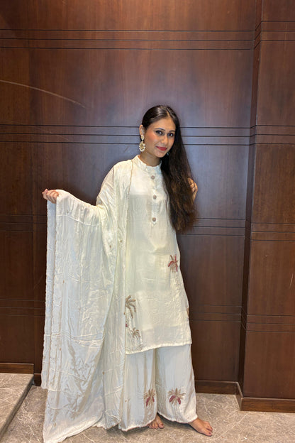 A woman stands barefoot in front of a dark wood-paneled wall, wearing a light cream/off-white traditional outfit with matching dupatta and embroidered details.