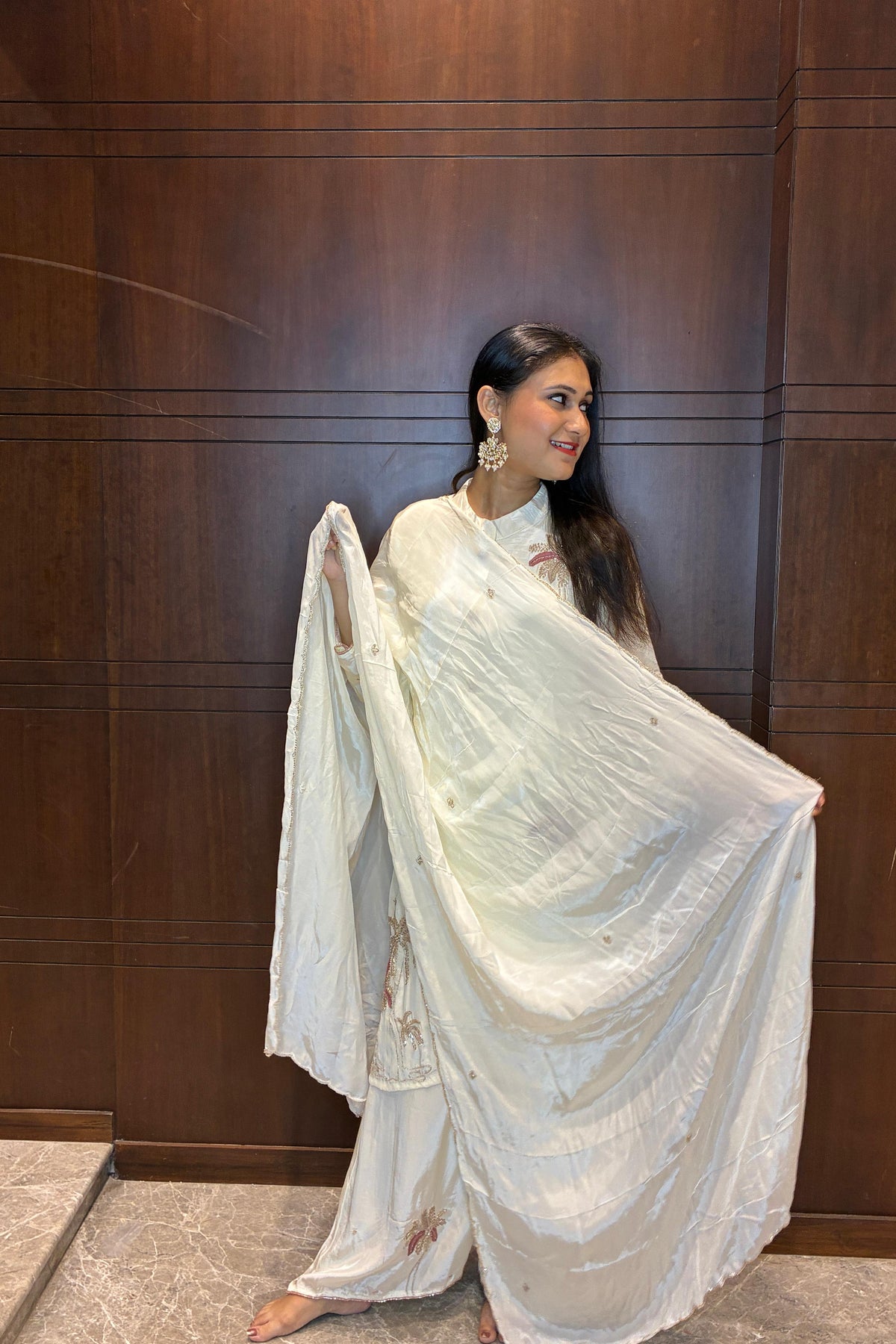 A woman wearing a white traditional outfit with a draped sari-style garment, standing against a dark wood-paneled wall and holding the fabric out to show its drape.