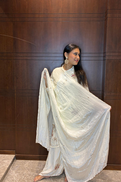 A woman wearing a white traditional outfit with a draped sari-style garment, standing against a dark wood-paneled wall and holding the fabric out to show its drape.