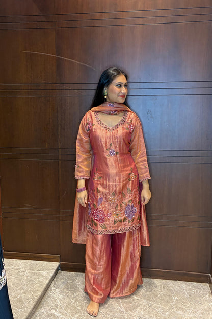 A woman stands barefoot indoors against a dark wood-paneled wall, wearing a peach and pink embroidered traditional outfit with matching pants and dupatta.