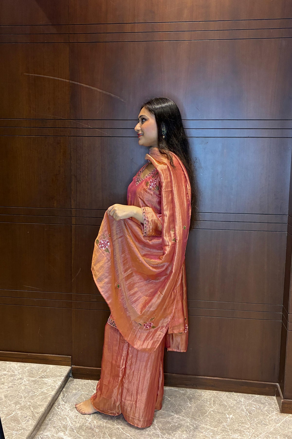 A woman stands in profile wearing a peach and orange embroidered traditional outfit with a matching dupatta, posed against a dark wood-paneled wall.
