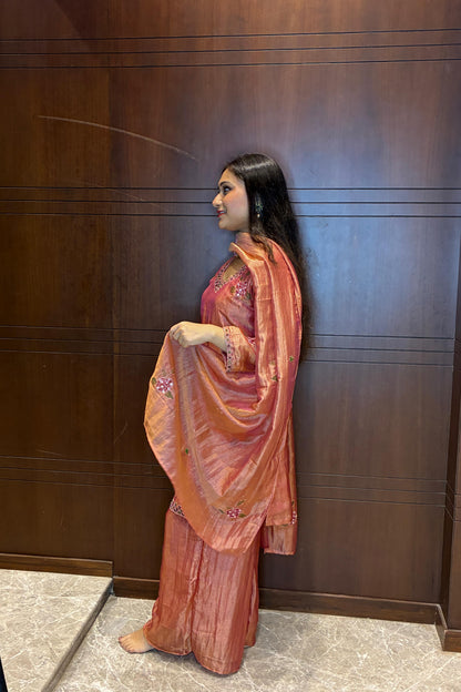 A woman stands in profile wearing a peach and orange embroidered traditional outfit with a matching dupatta, posed against a dark wood-paneled wall.