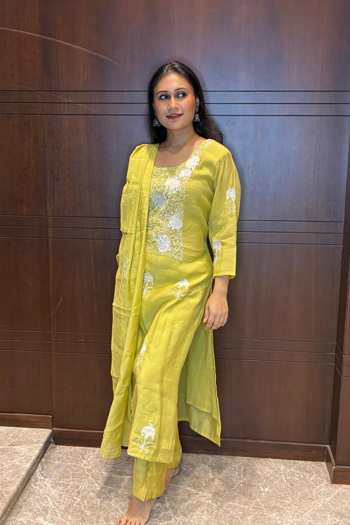 A woman wearing a bright yellow-green embroidered kurta set with a matching dupatta, standing against a dark wood-paneled wall.