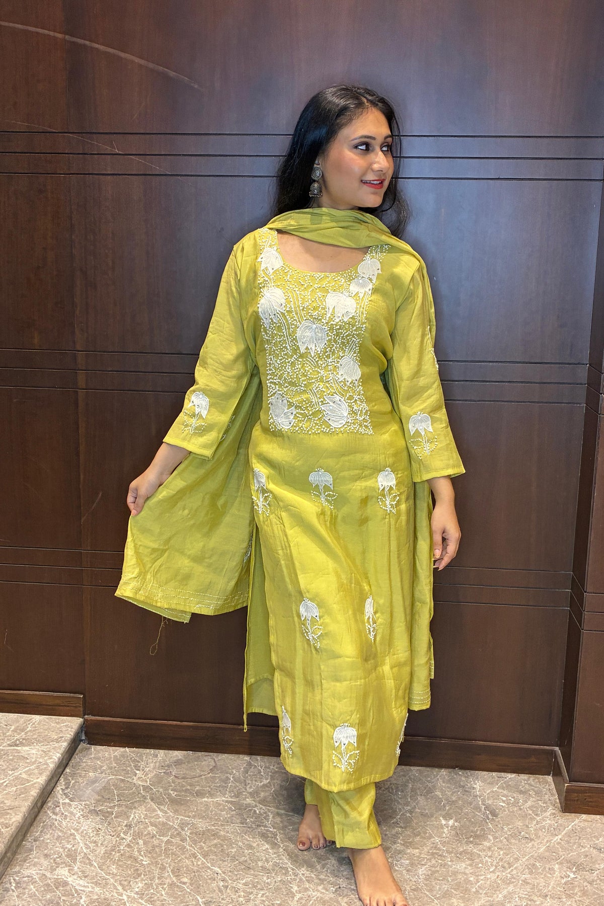 A woman modeling a lime green traditional Indian salwar kameez suit with white floral embroidery, standing against a dark wood-paneled wall.