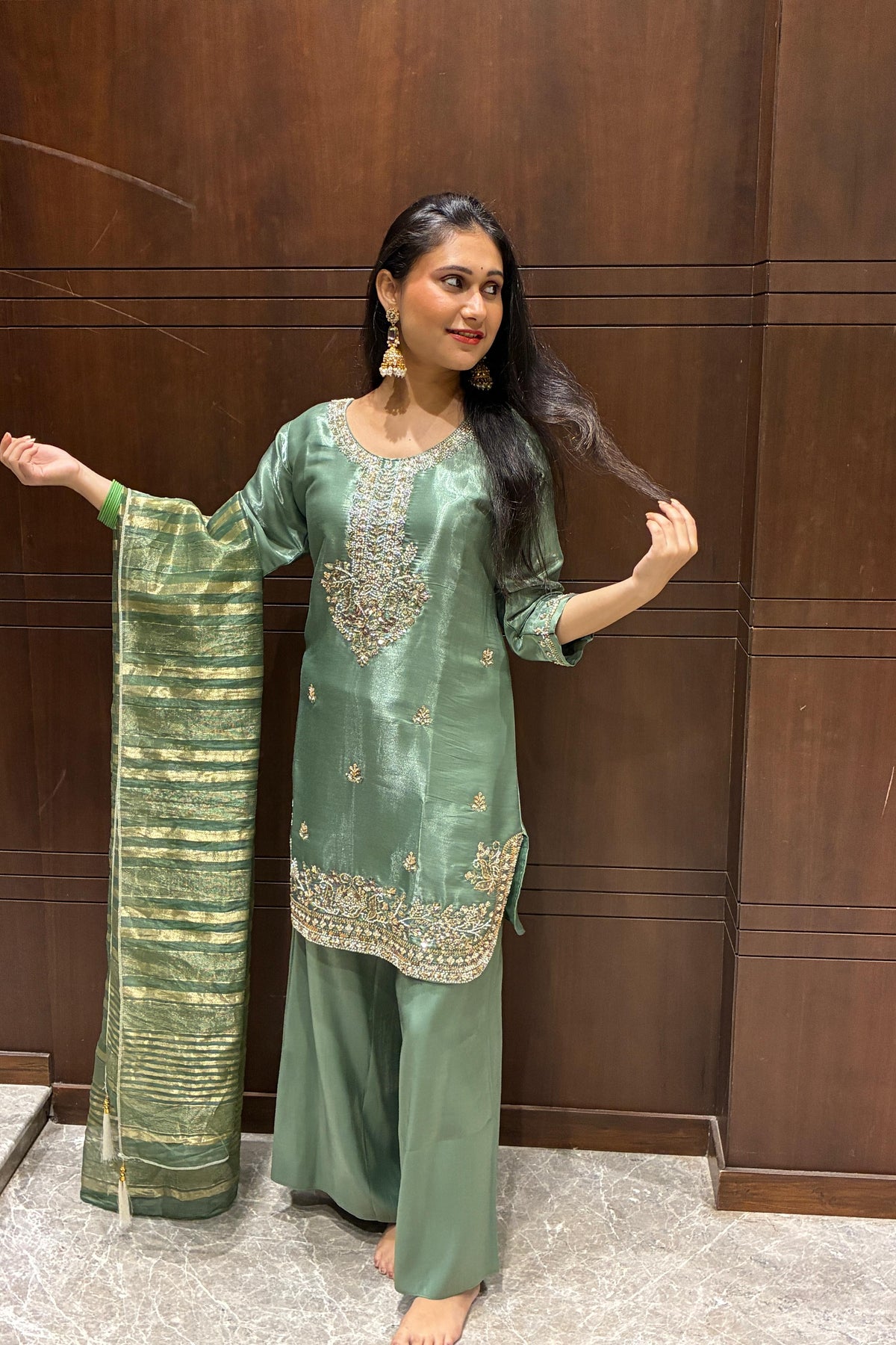 A woman modeling a sage green three-piece salwar kameez suit with gold embroidery and a matching striped green dupatta, standing in front of a dark wood-paneled wall.