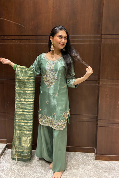 A woman modeling a sage green three-piece salwar kameez suit with gold embroidery and a matching striped green dupatta, standing in front of a dark wood-paneled wall.