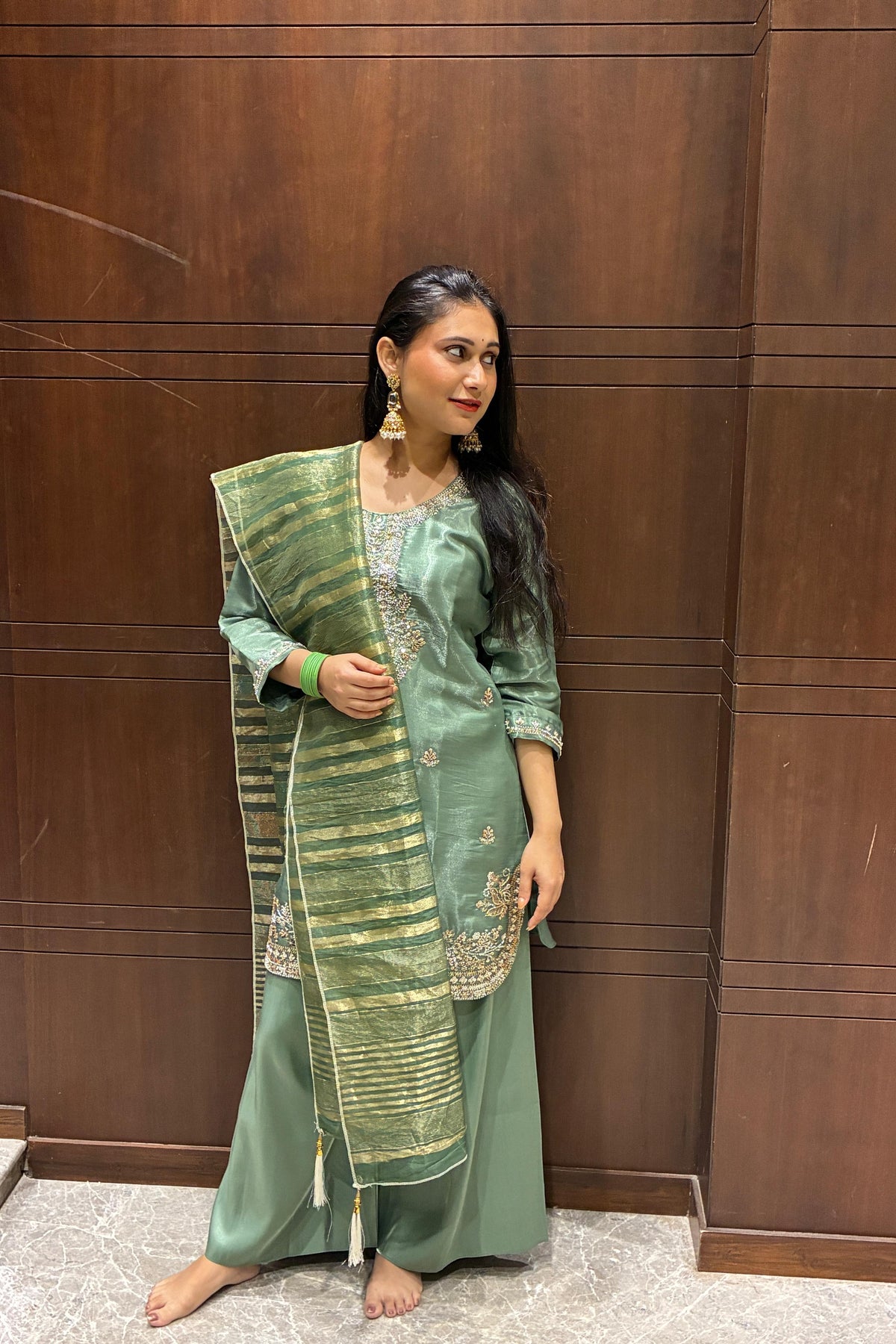 A woman stands barefoot in front of a dark wood-paneled wall, wearing a sage green traditional outfit with gold embroidery and a matching patterned dupatta.