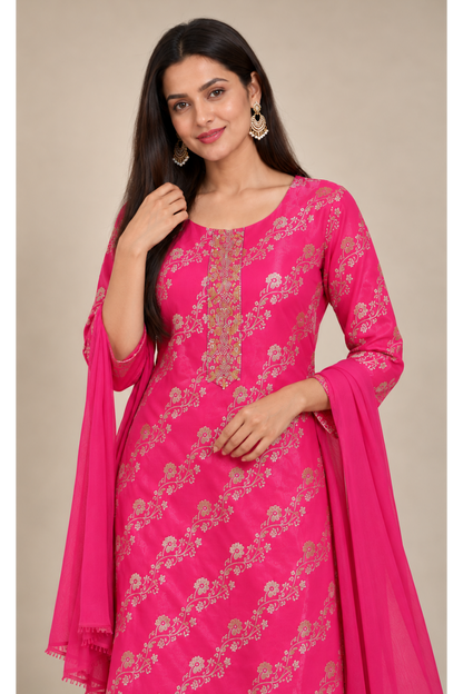 A woman modeling a bright pink traditional Indian salwar kameez set with gold floral embroidery and a matching pink dupatta, set against a plain beige background.