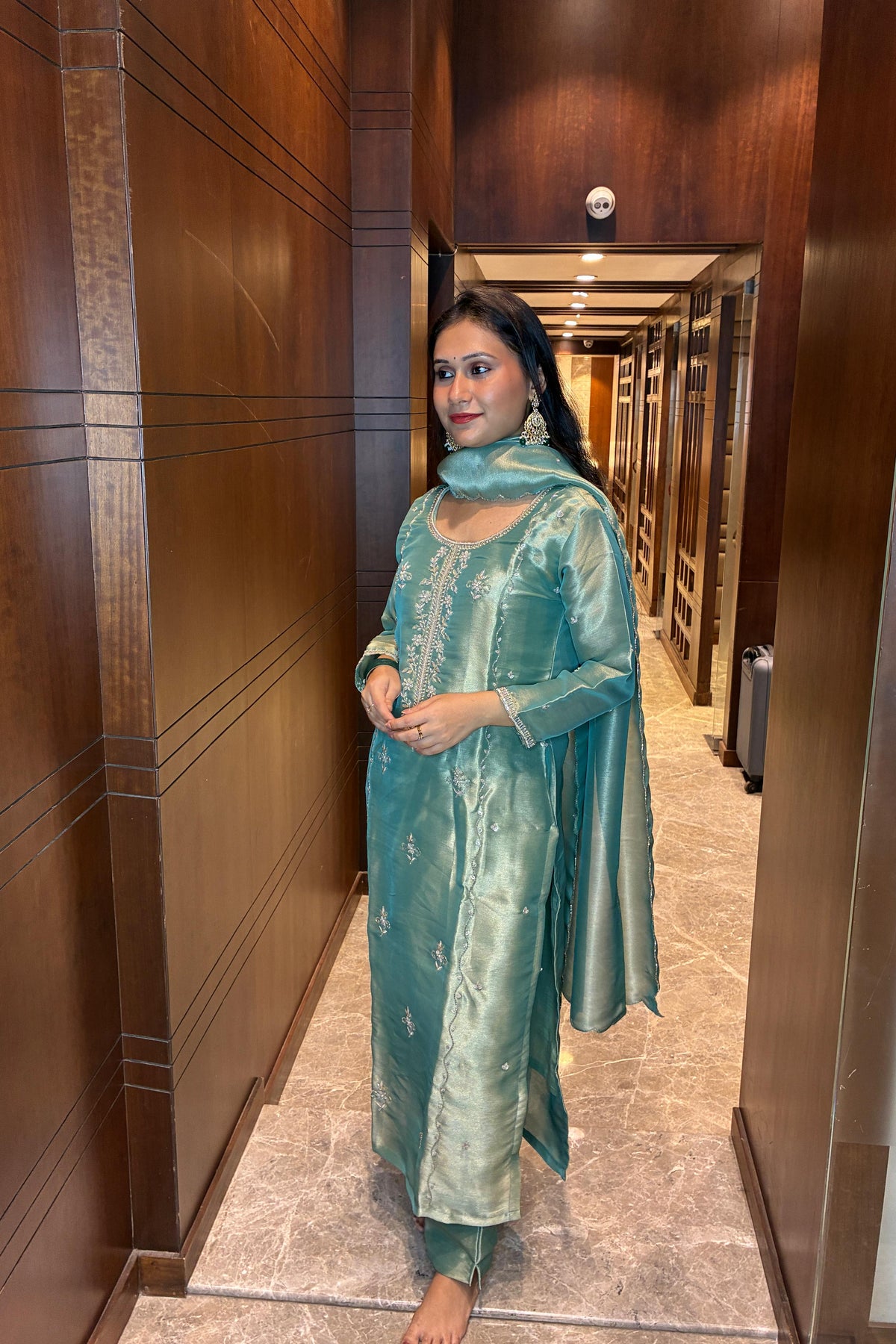 A woman wearing a metallic teal green traditional Indian salwar kameez suit with a matching dupatta, standing in a hallway with dark wood-paneled walls.