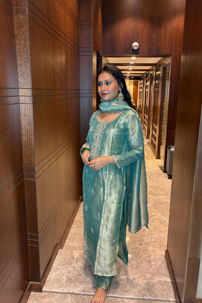 A woman wearing a metallic teal green traditional Indian salwar kameez suit with a matching dupatta, standing in a hallway with dark wood-paneled walls.