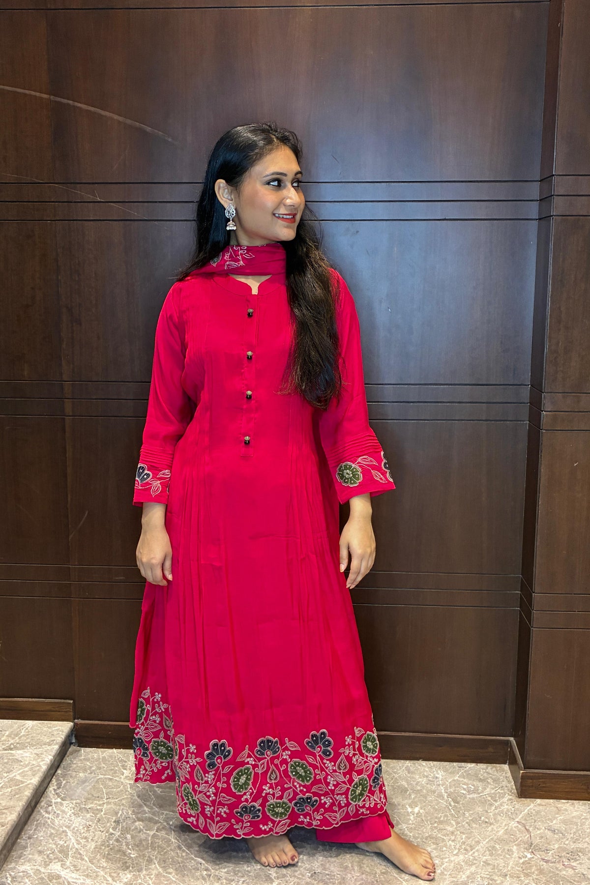 A woman modeling a bright pink traditional long kurta with green and gold floral embroidery on the hem and cuffs, standing against a dark wood-paneled wall.