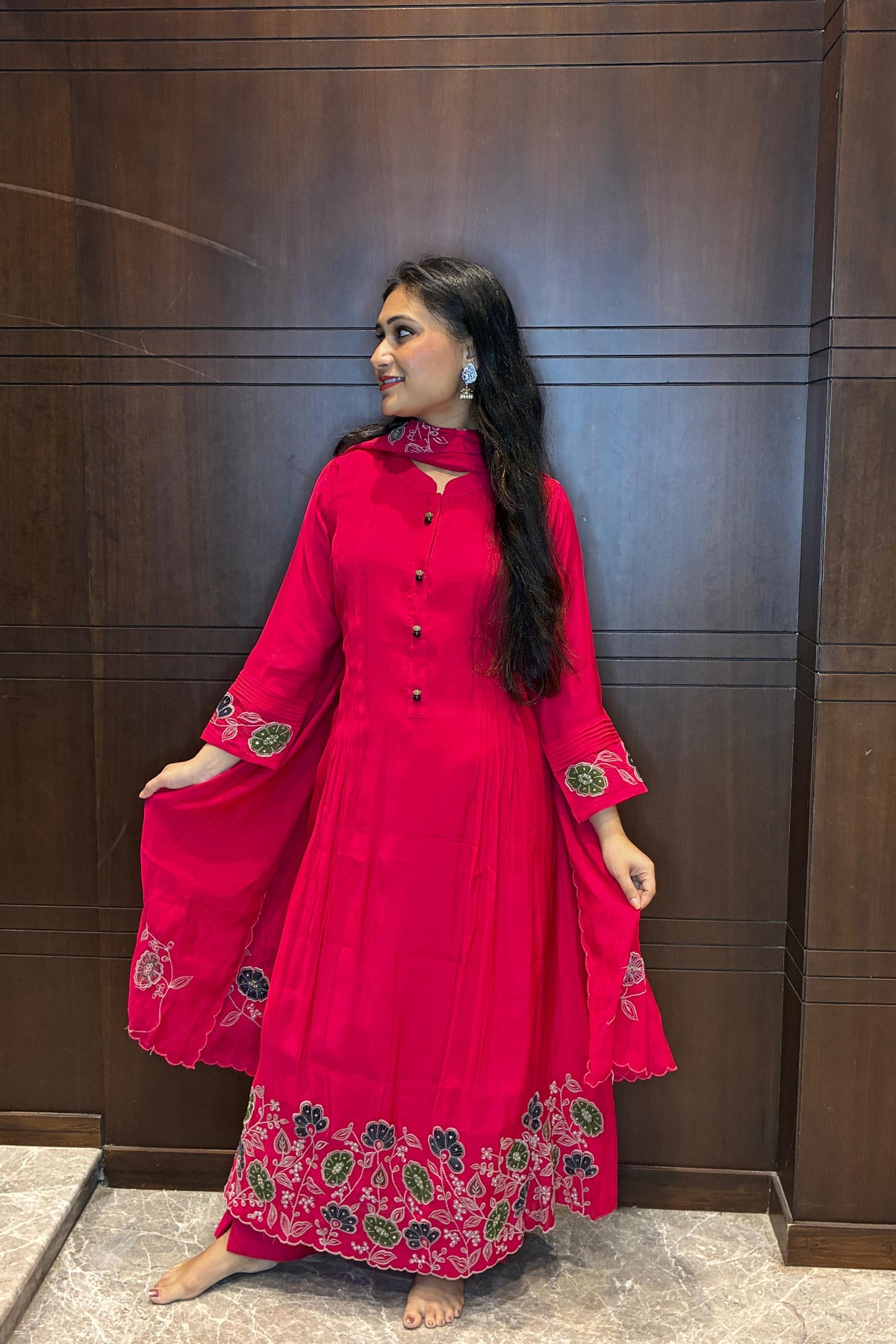 A woman wearing a bright pink traditional long dress with green and gold floral embroidery, standing in front of a dark wood-paneled wall.