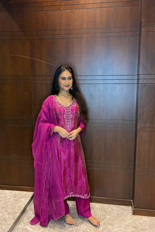 A woman wearing a vibrant magenta pink traditional Indian outfit with matching dupatta and trousers, standing in front of a dark brown wooden paneled wall.