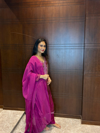 A woman wearing a bright magenta pink traditional outfit with a matching dupatta, standing in front of a dark brown wooden paneled wall.