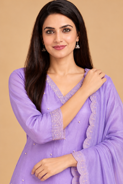 A woman wearing a lavender purple traditional outfit with matching sheer dupatta, featuring scalloped embroidered borders and small bead embellishments, set against a solid beige background.